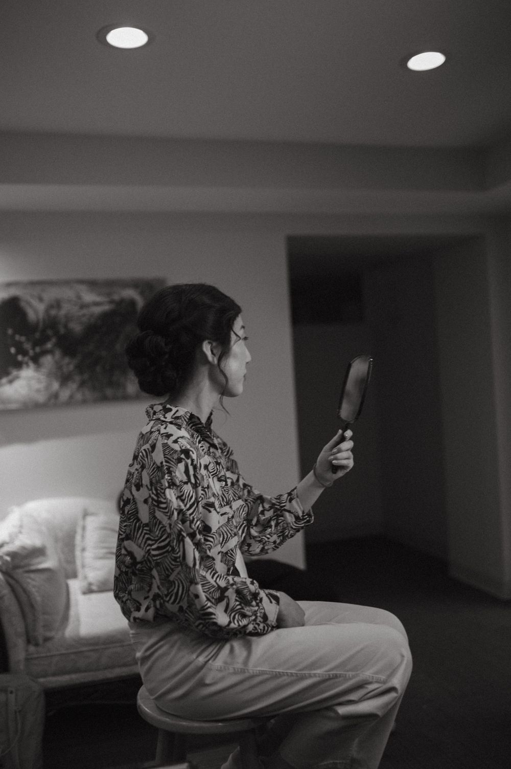A person in a patterned dress holds a mirror while getting ready in a dimly lit room.