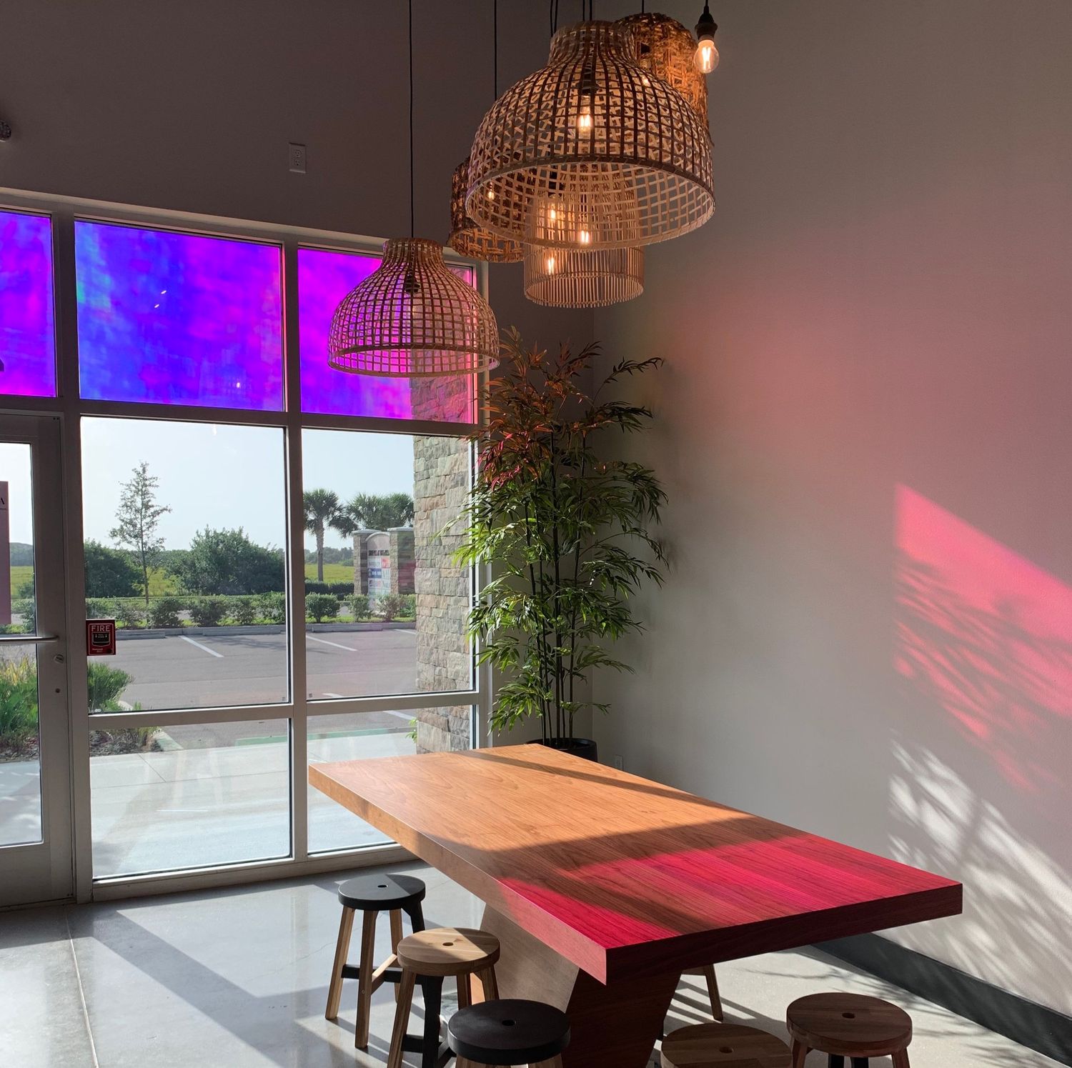 Modern cafe interior with wicker pendant lights, wooden table, and vibrant purple-lit windows.