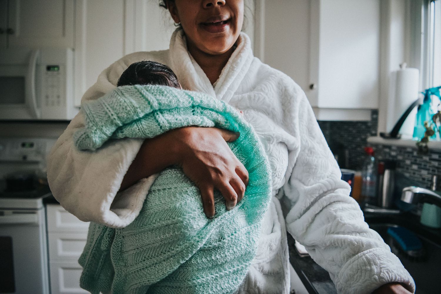 A newborn is swaddled up in a handmade knit light teal blanket as he's held by his mother in a fluffy white housecoat.