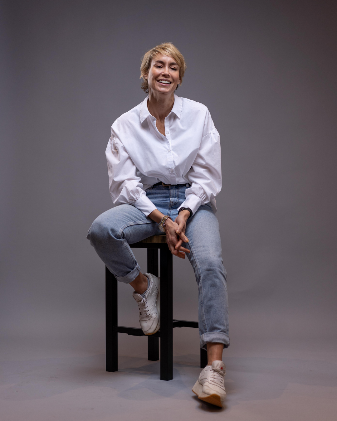 Image of a classic model photoshoot in the photography studio. She's sat on a stool wearing a white shirt and blue jeans
