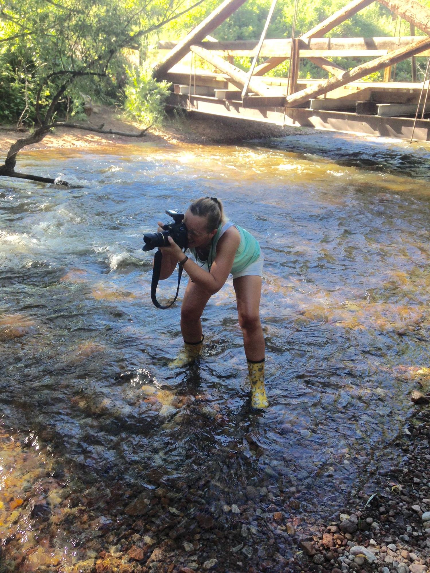 Photographer in shorts and boots stands in flowing stream while taking pictures.