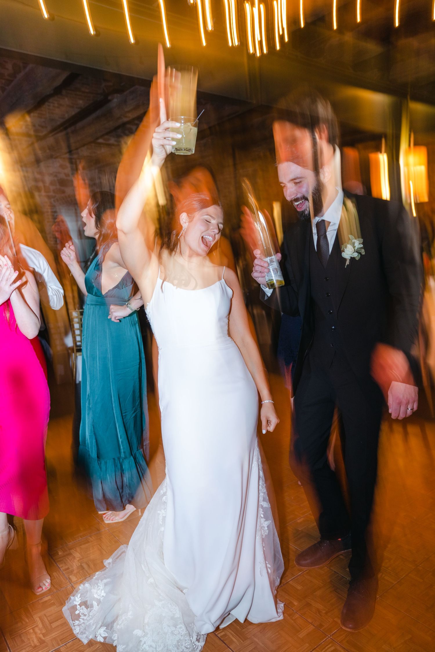 Bride and groom dancing joyfully with guests at a lively wedding reception under warm string lights.