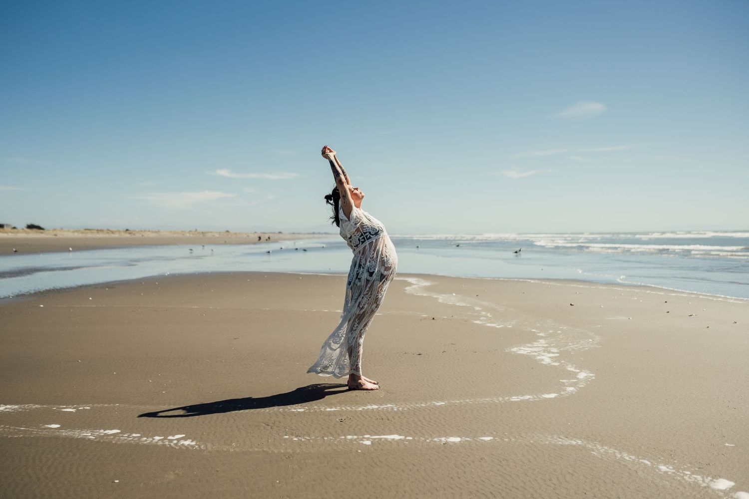 A person in flowing clothes dances joyfully on a beach against a blue sky.