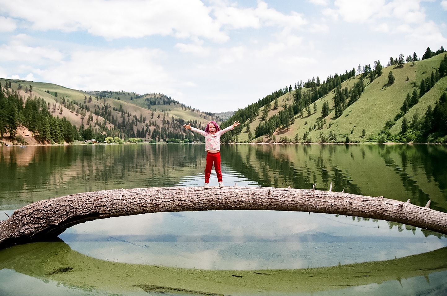 Person with outstretched arms stands on fallen log over mountain lake.