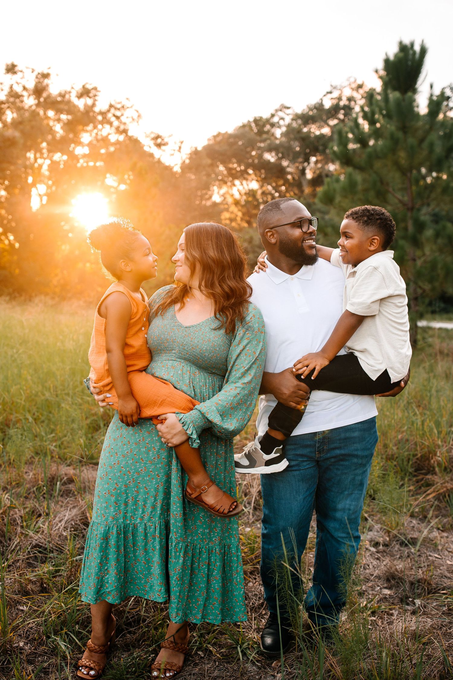 Happy interracial family posing together at golden hour in a field, parents holding two young children.