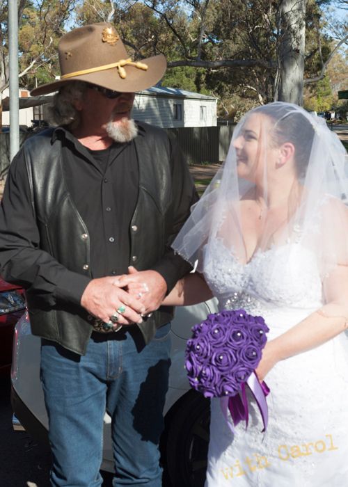 Wedding couple shares intimate moment while holding purple bouquet outdoors.