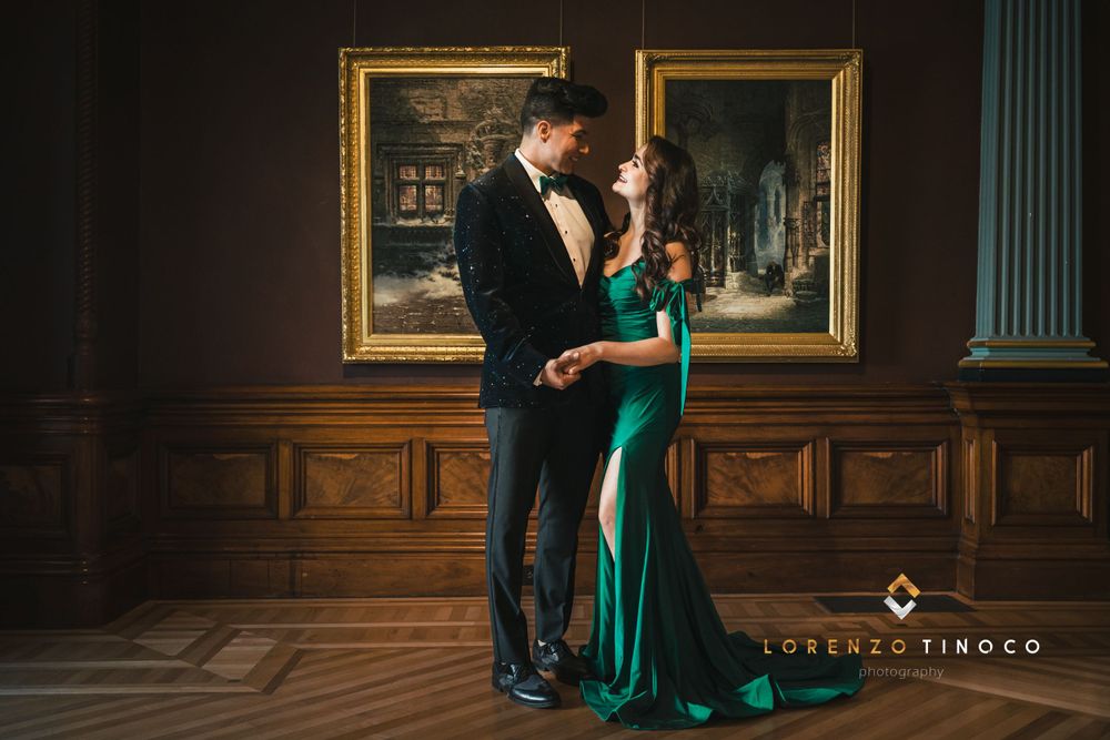 A glamorous couple in formal attire poses against ornate wood paneling and framed artwork.