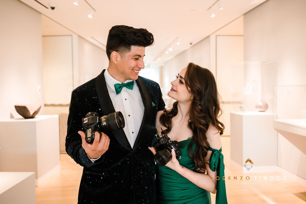 A couple in formal attire shares a playful moment while holding vintage cameras in an art gallery.
