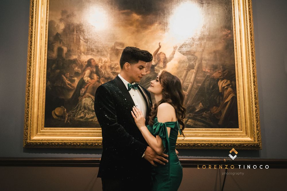 A formal couple poses intimately in front of a dramatic historical painting in a museum setting.
