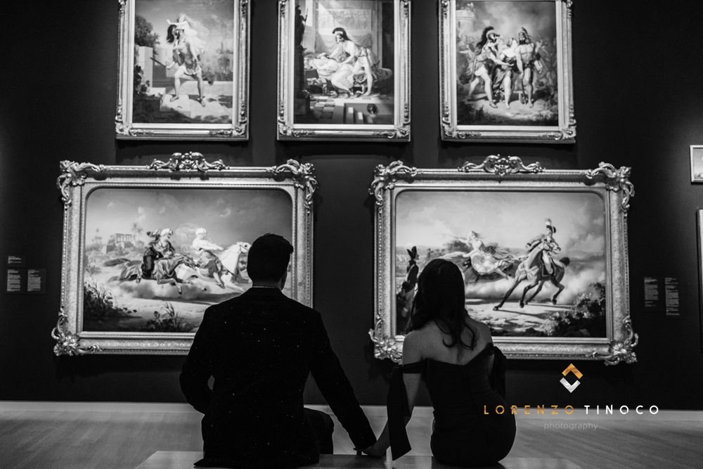 A silhouetted couple admires framed classical paintings in a dimly lit museum gallery.