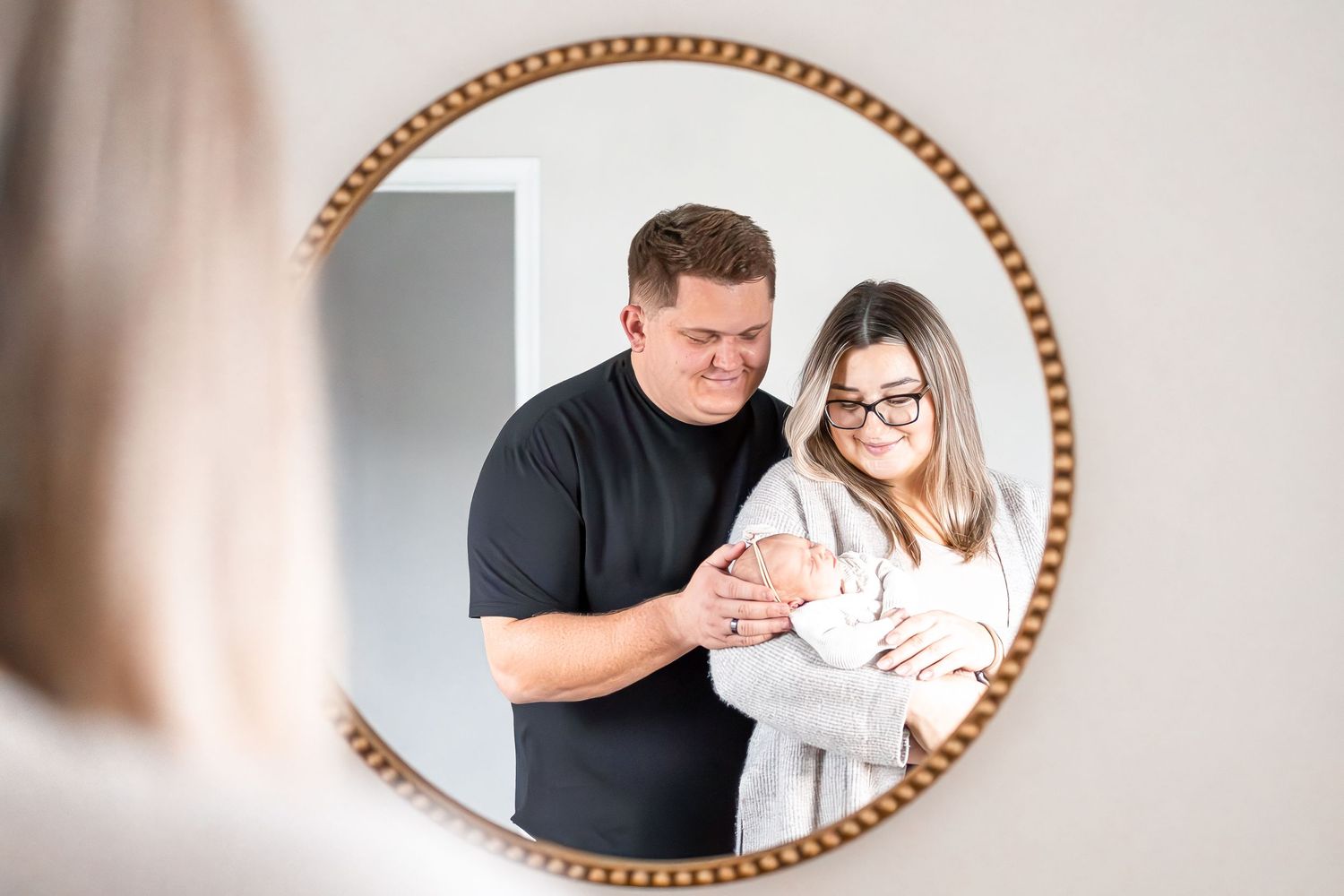 A family stands together looking at their newborn baby, reflected in a decorative round mirror.