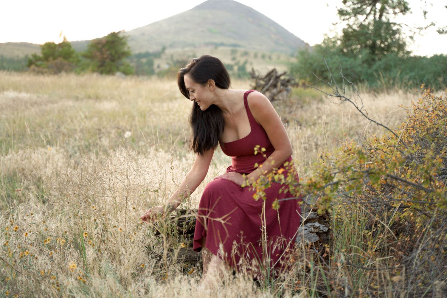 Figure in burgundy dress kneeling in wildflower meadow with mountain backdrop.