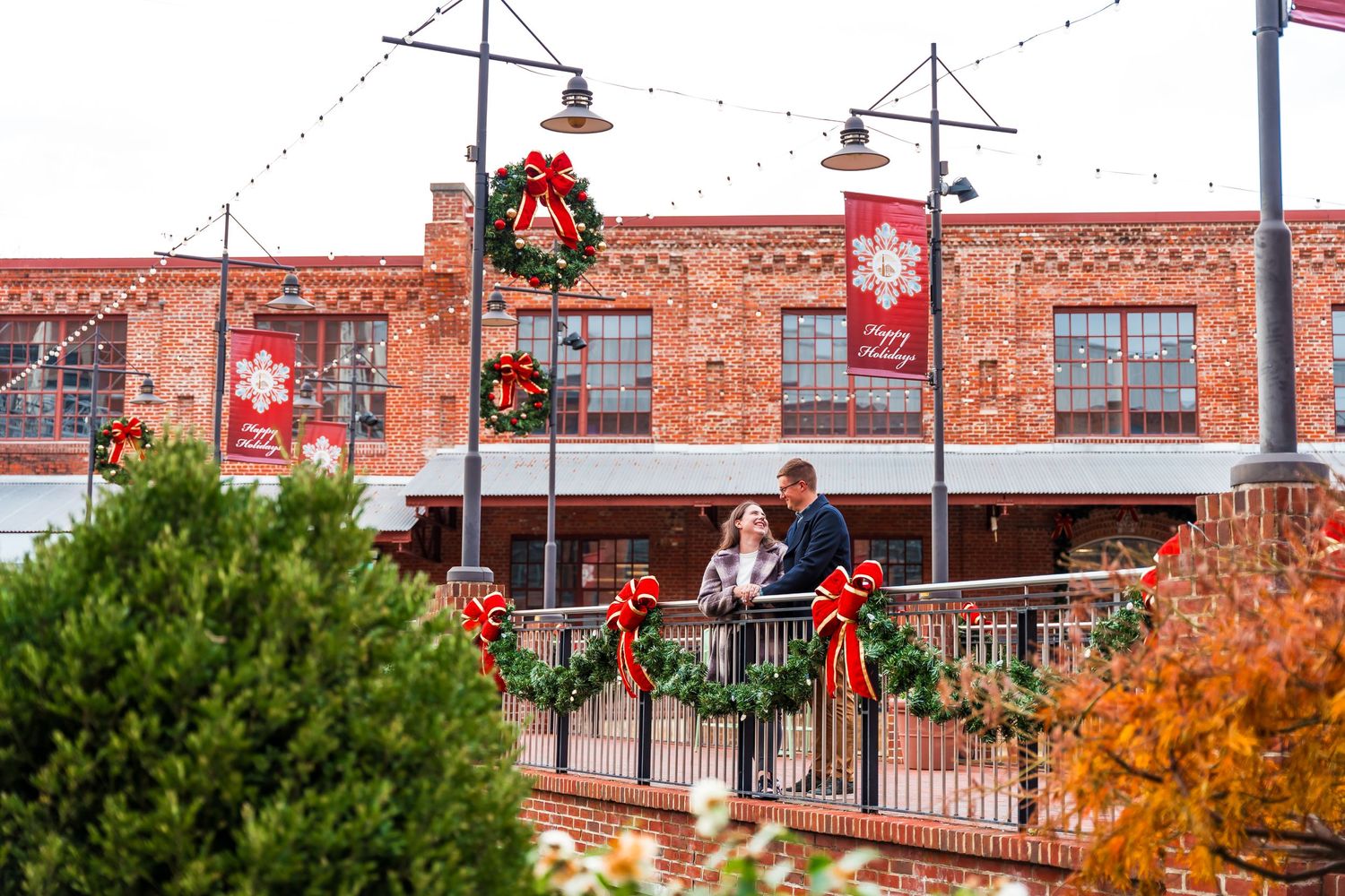 Couple shares a romantic moment on a decorated balcony surrounded by Christmas wreaths and festive red banners.