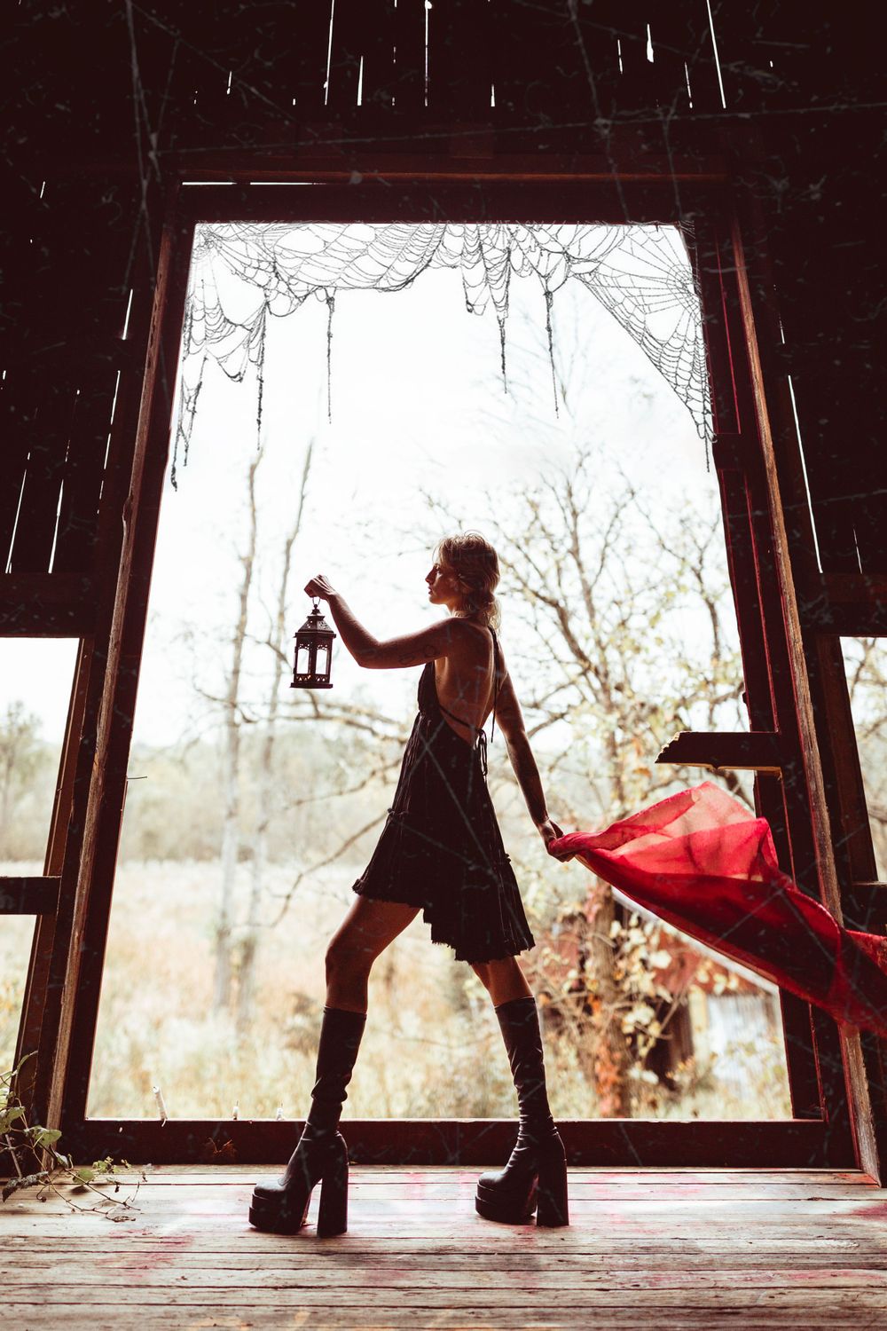Silhouette holding lantern in doorway wearing platform boots with flowing red fabric creating dramatic mood.