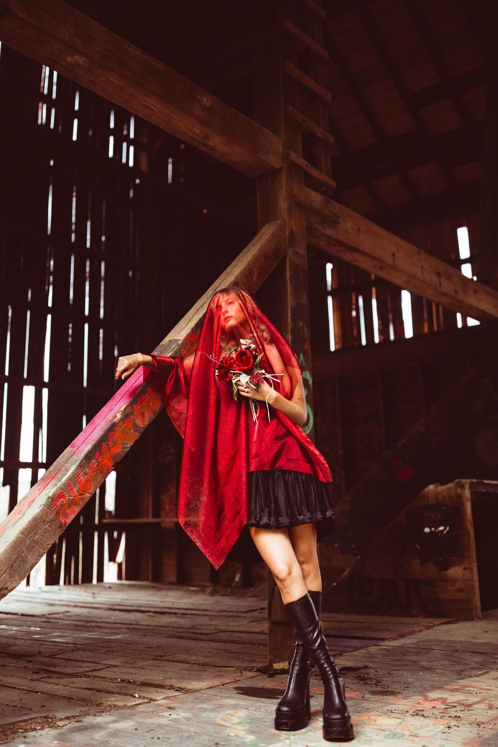 Figure in red cape and platform boots poses dramatically in rustic barn setting.