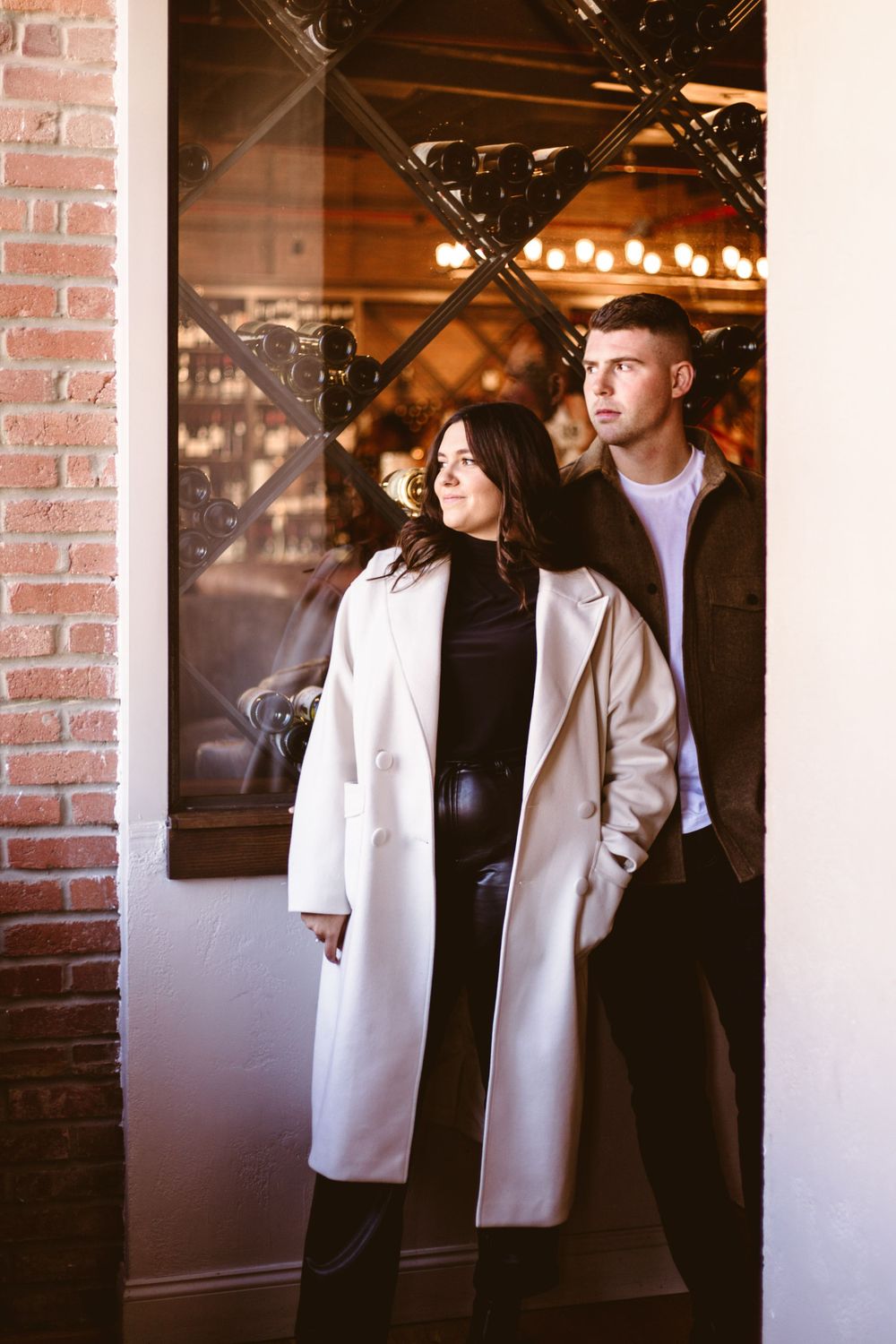 Stylish couple poses by brick wall in urban setting with warm lighting.