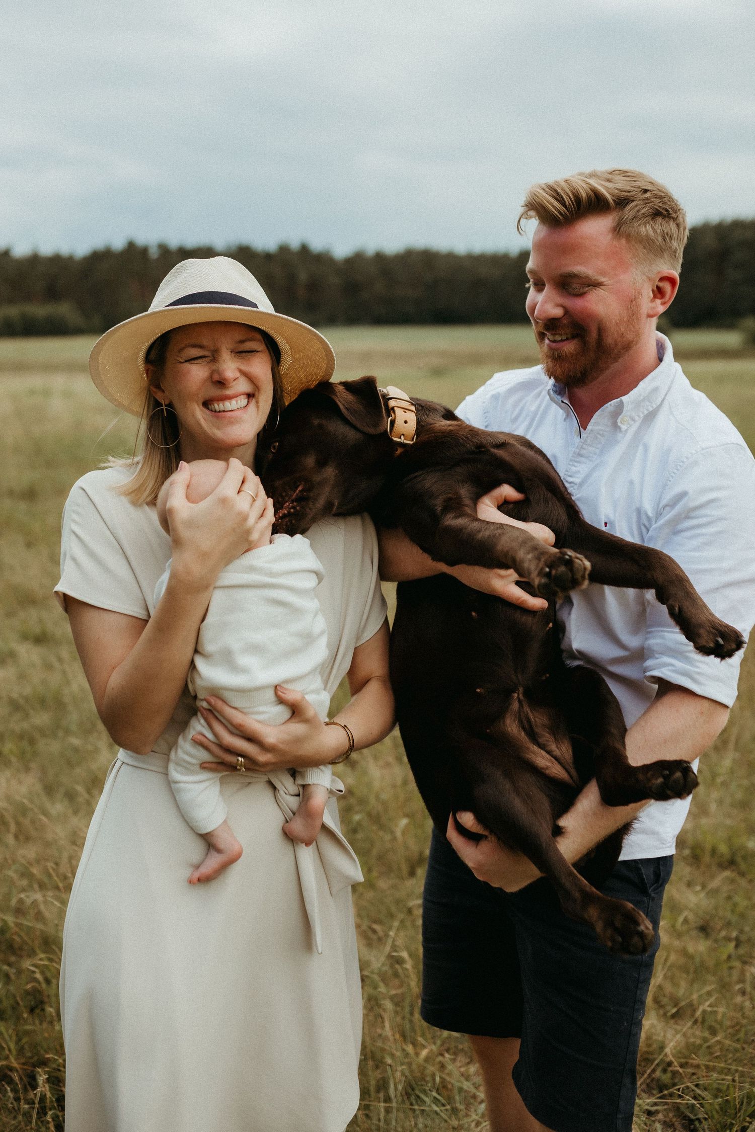 Couple with their chocolate lab dog posing together in a sunny meadow.