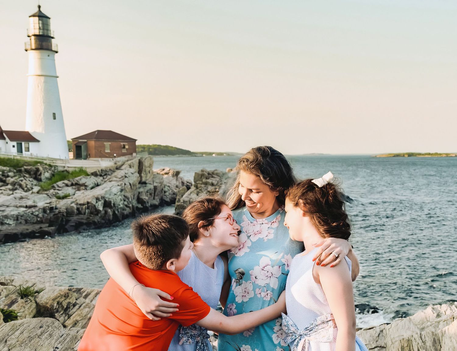A mother smiles at her three children while posing by the ocean and the Portland Head Light Lighthouse at Fort Williams.