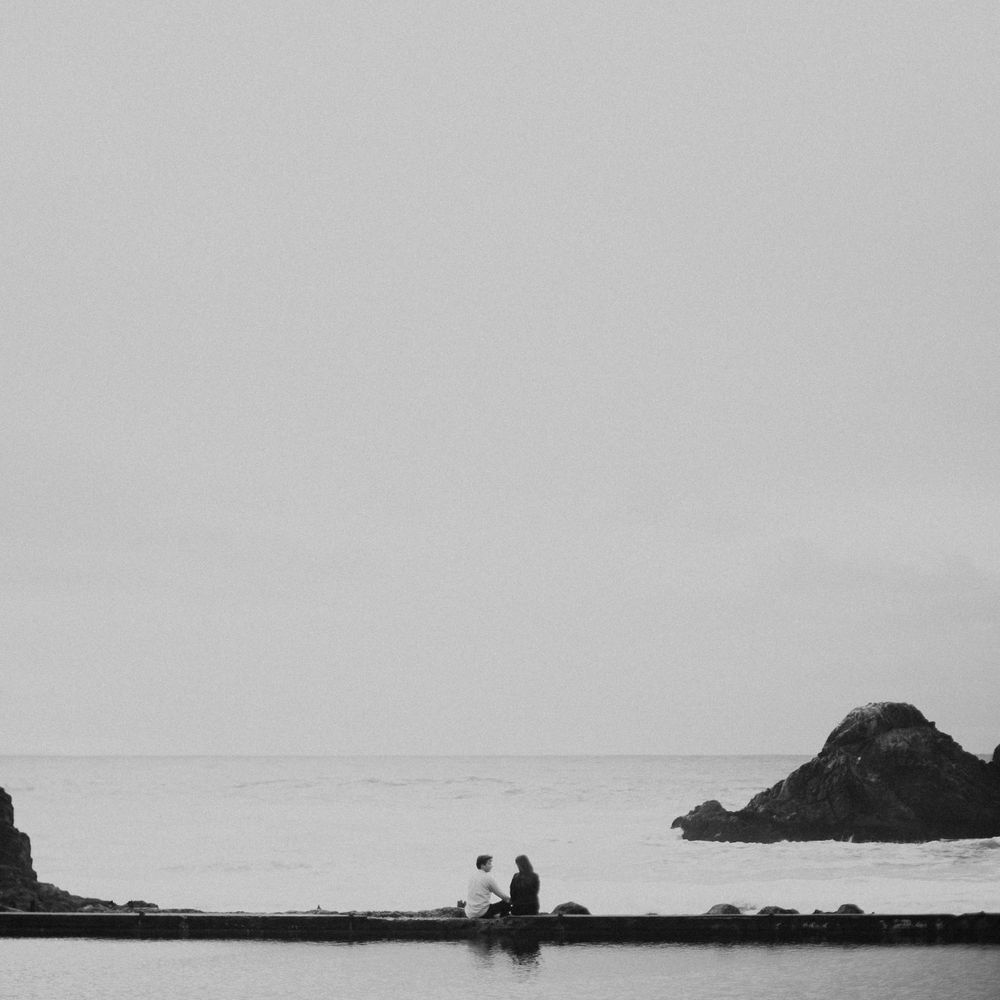 Two silhouettes sit together on a rocky outcrop overlooking a tranquil ocean horizon at dusk.