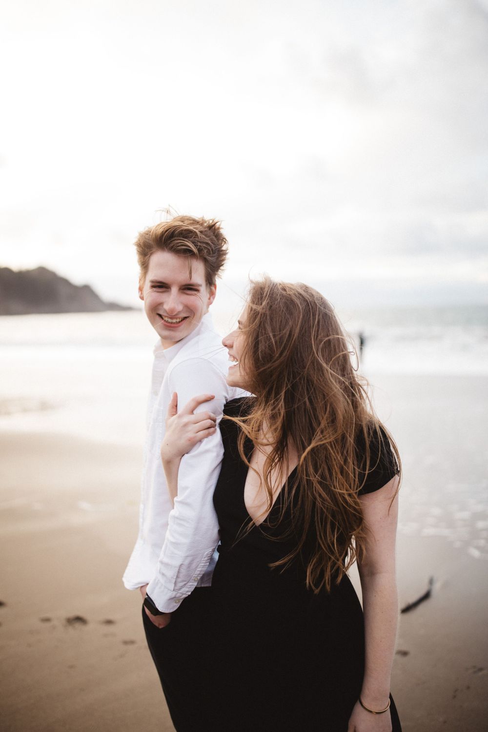 A joyful couple shares a candid moment while walking along a misty beach at sunset.