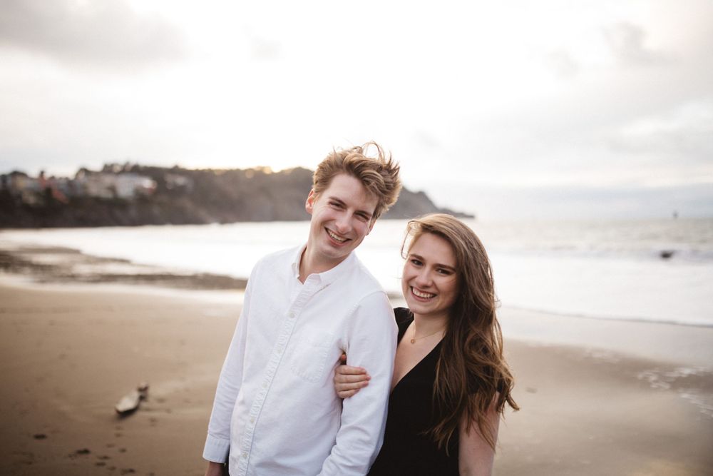 A couple shares a cheerful moment together on a cloudy beach with coastal cliffs in the distance.