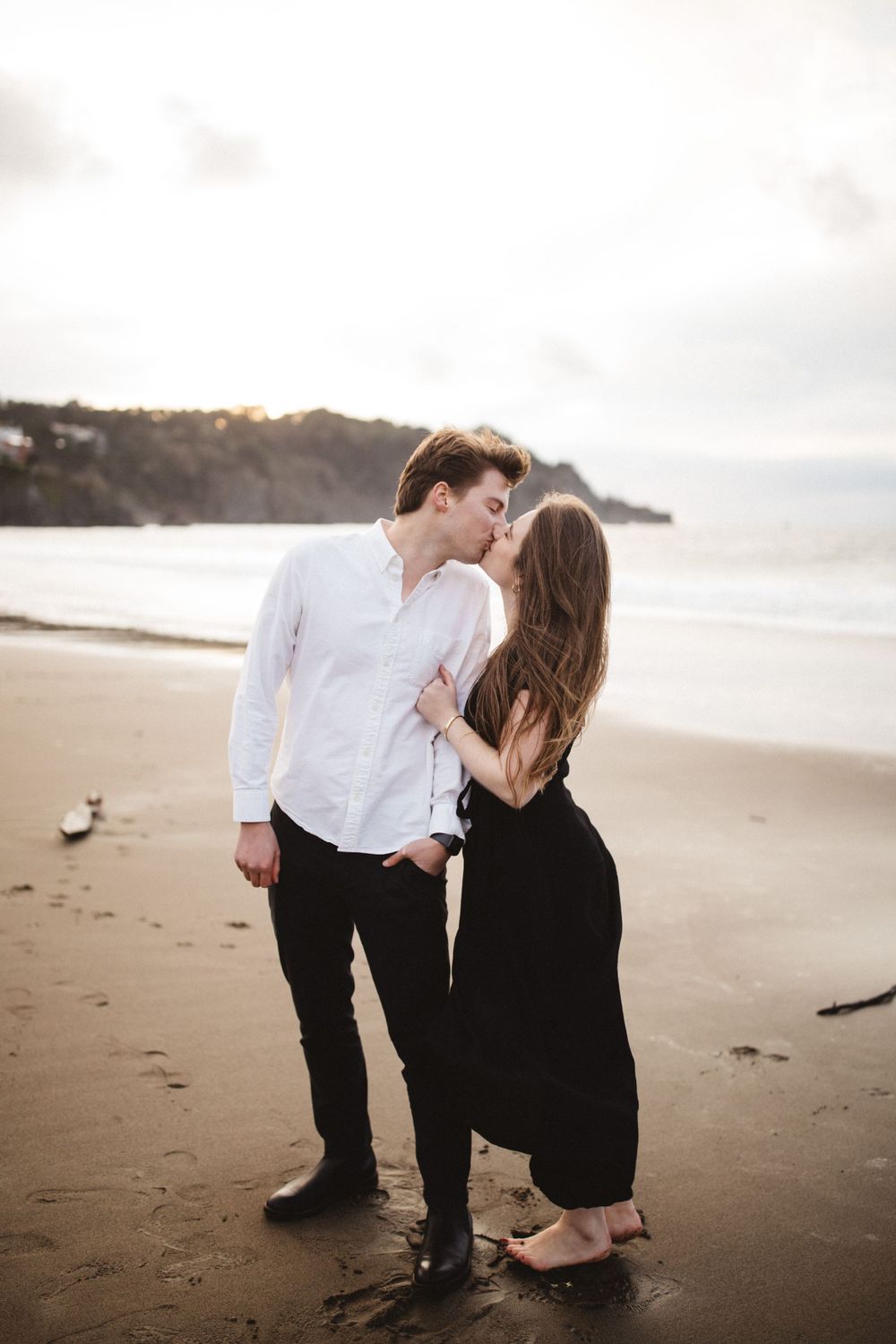 A romantic couple embraces on a secluded beach with cliffs and ocean waves in the background.
