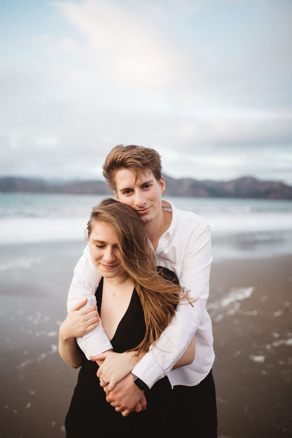 A tender embrace between a couple on a cloudy beach with waves crashing behind them.