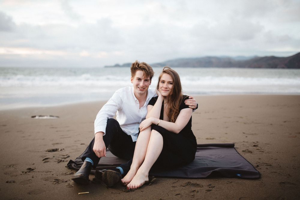 A couple relaxes together on a blanket at the beach with mountains visible in the distance.