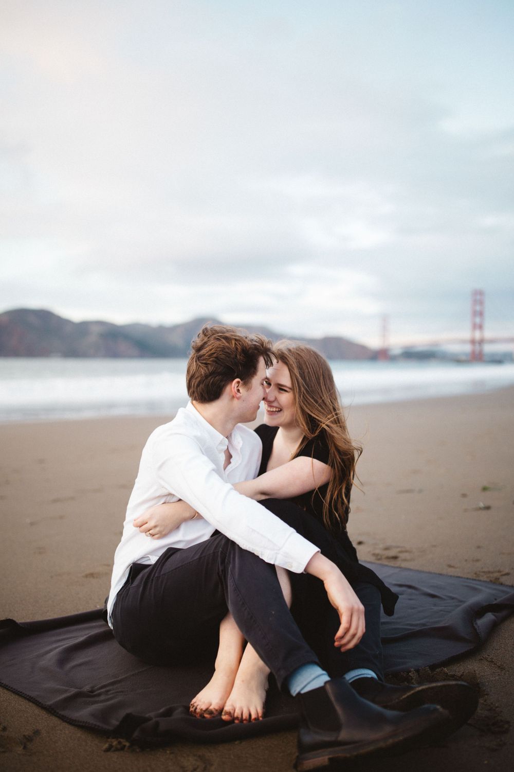 A couple shares an intimate moment on a blanket at Baker Beach with the Golden Gate Bridge visible.