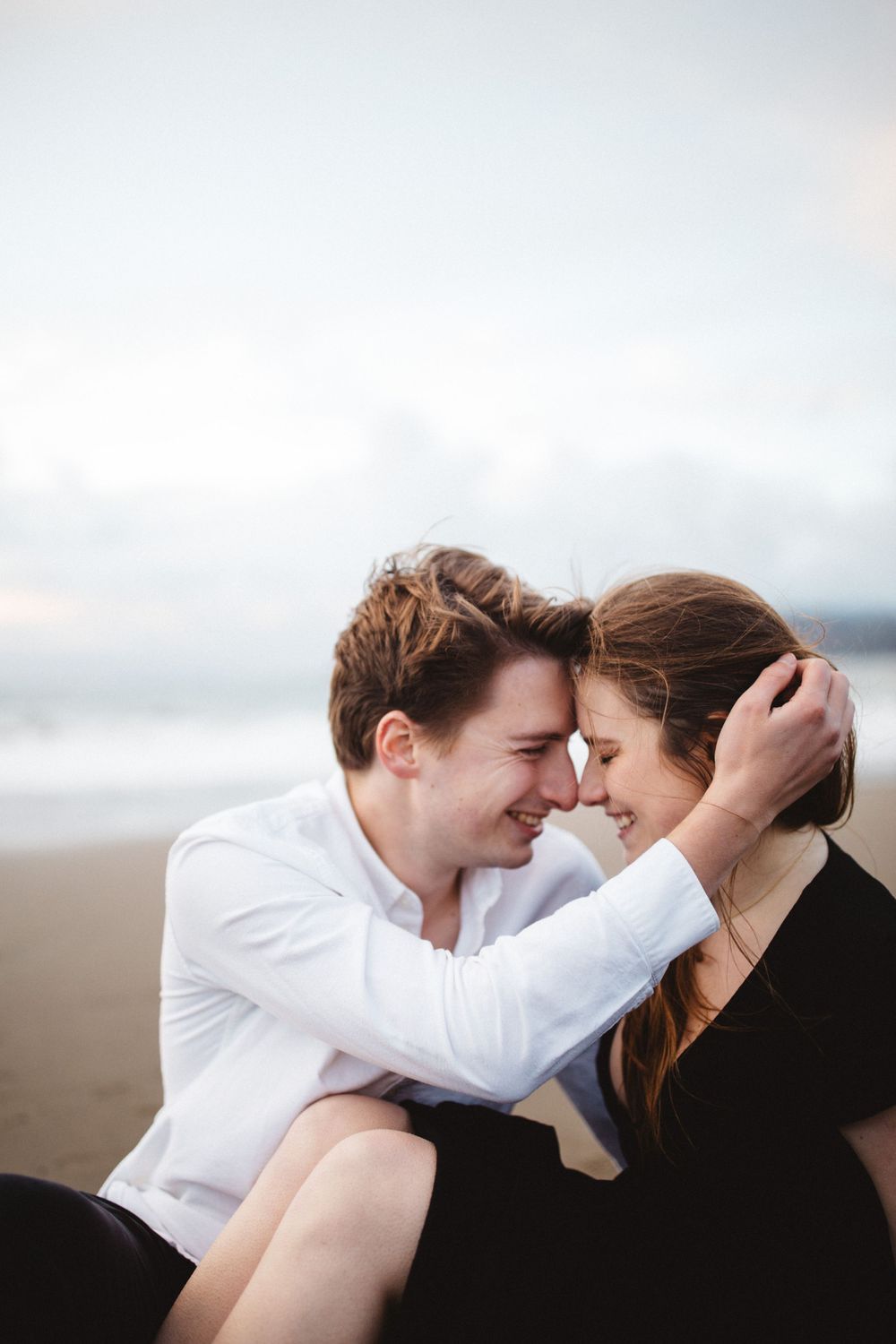 A joyful couple shares an intimate moment while sitting close together on a cloudy beach.