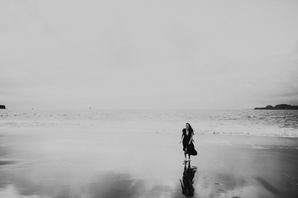A silhouetted figure stands alone on a reflective beach shoreline in black and white.