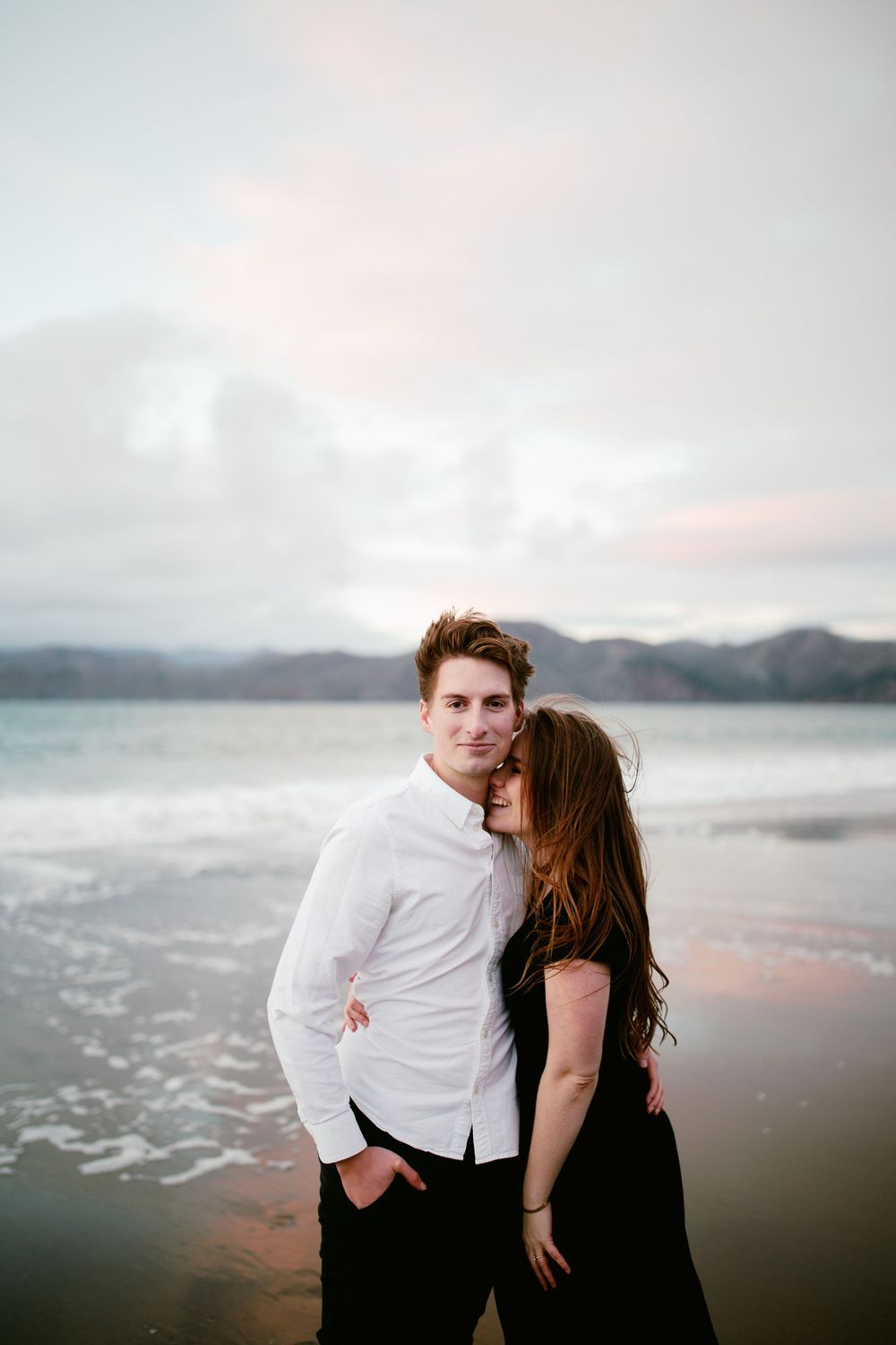 A romantic couple stands close together on a misty beach at sunset with coastal mountains in the distance.
