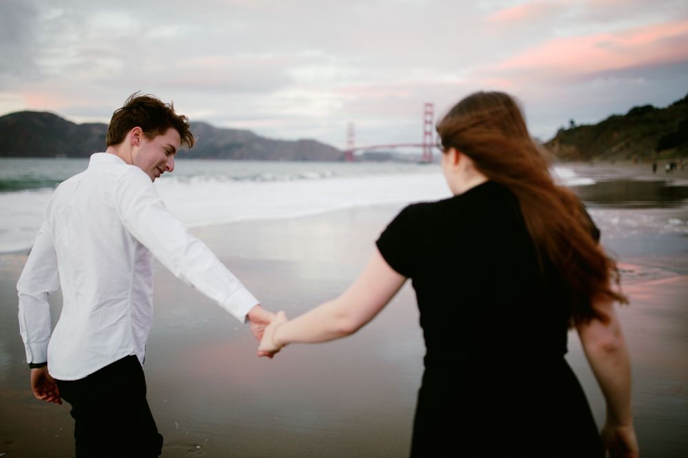 A couple holding hands walking on a beach at sunset with mountains in the background.