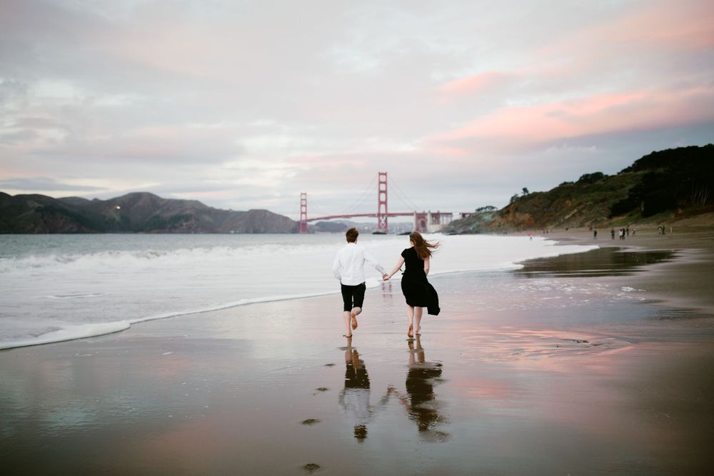 Two people walk along a beach at sunset with the Golden Gate Bridge visible in the distance.