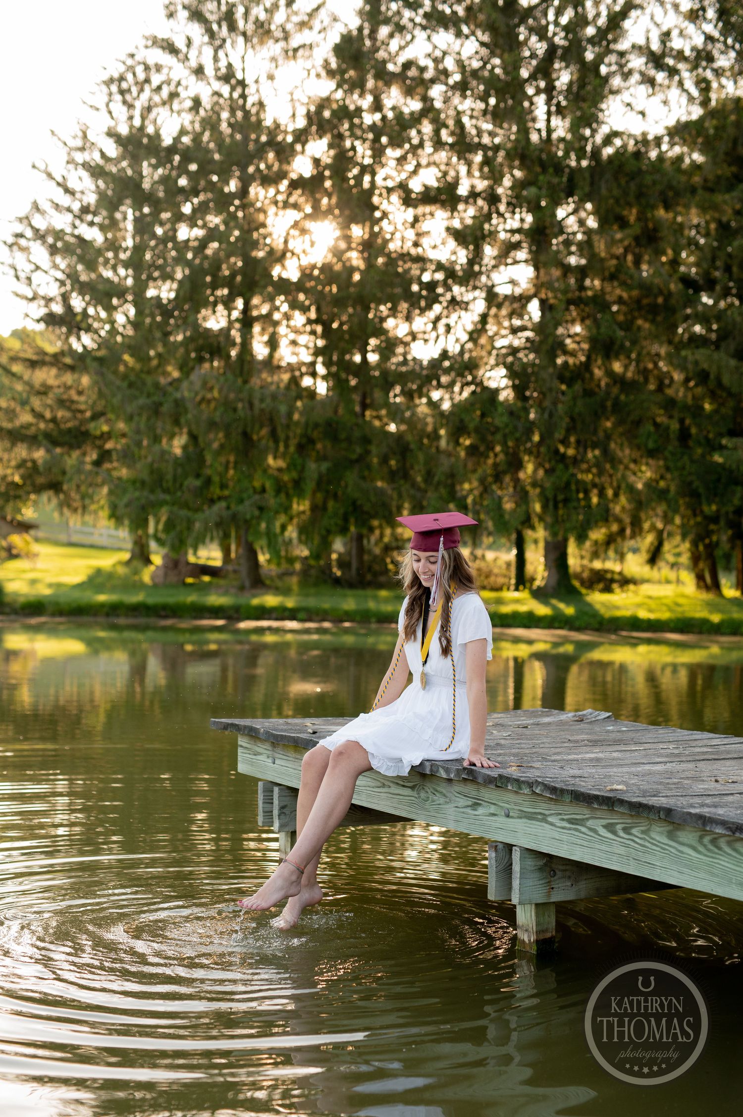 Graduate dips feet in pond while sitting on wooden dock during golden hour.