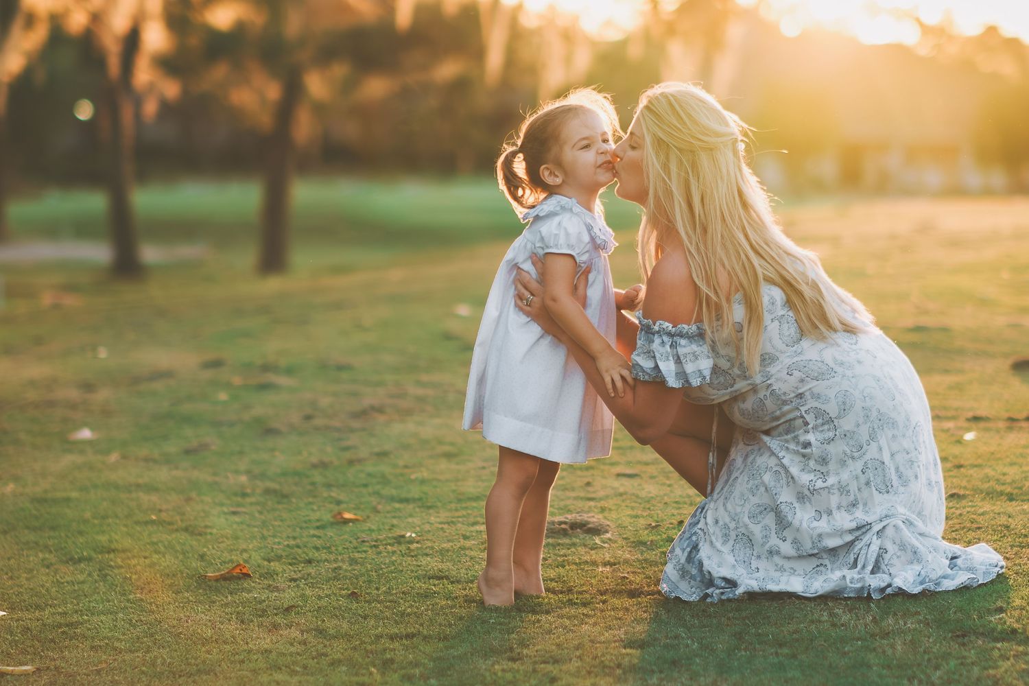 Touching moment at sunset in a grassy field with white lace dress and golden sunlight streaming through trees.