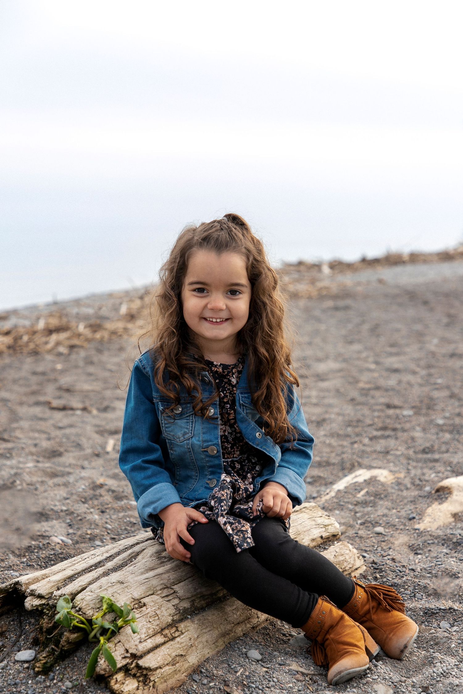 A young girl sitting on a log at the beach wearing a jean jacket