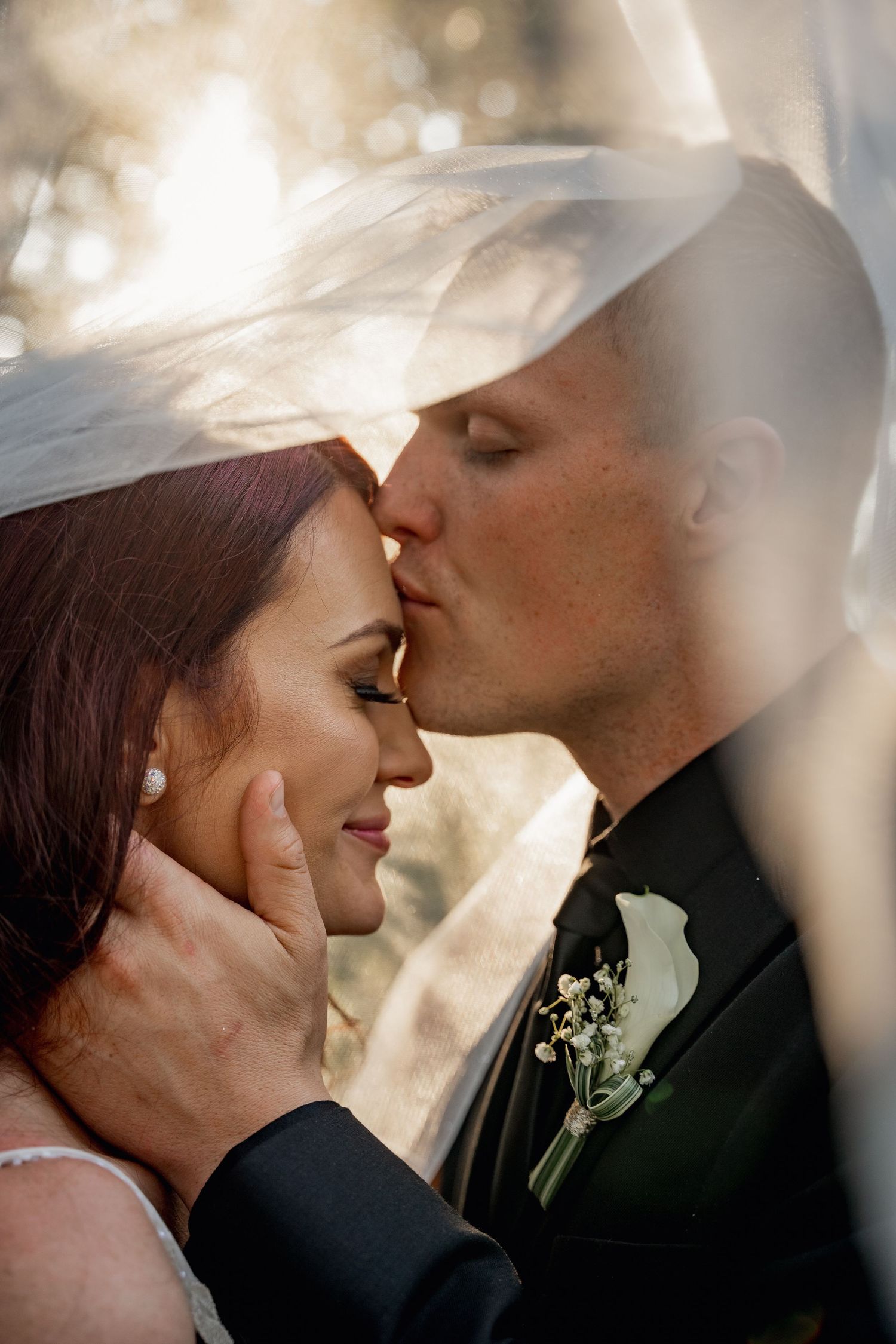 A romantic wedding moment captured through delicate veil in soft sunlight with a boutonniere visible on the groom's lapel.