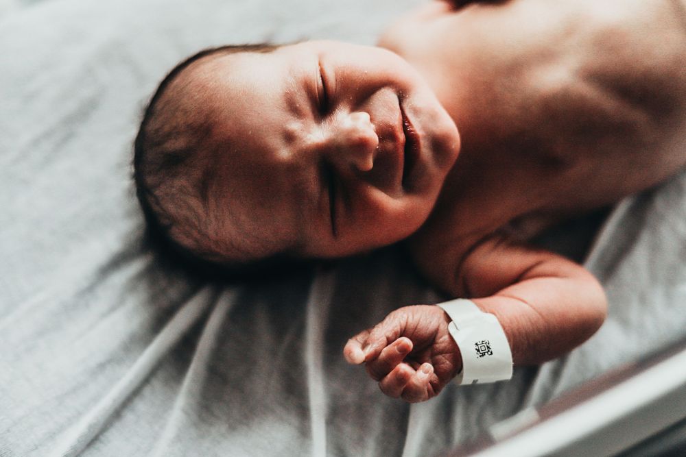 A newborn baby wearing a hospital bracelet lies on light gray bedding while crying. terre haute family wedding photographer parke county
