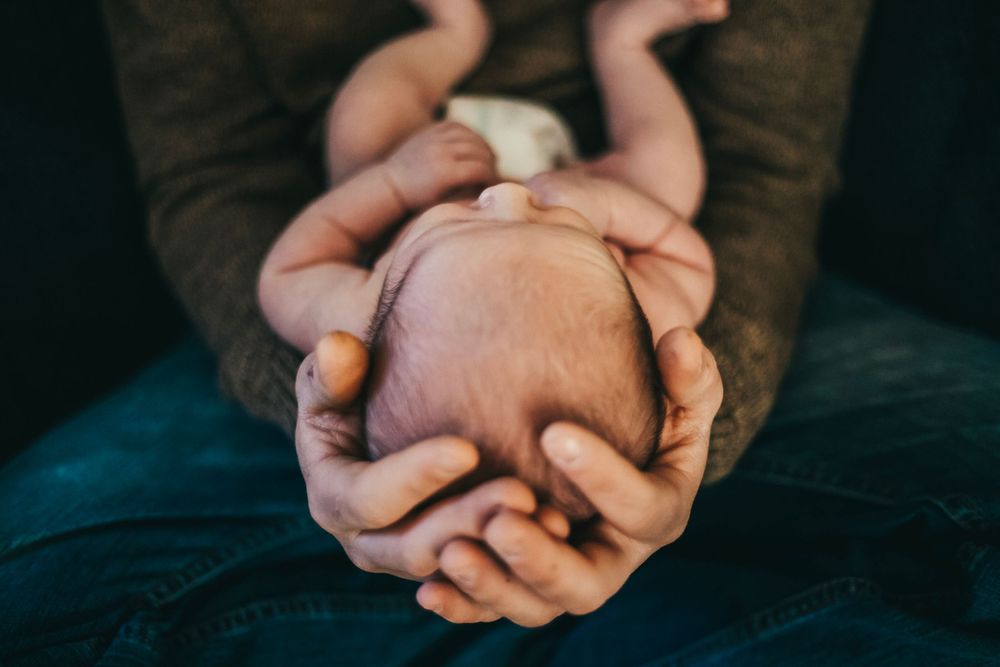 Two sets of hands tenderly hold and support a tiny sleeping newborn baby on a dark fabric background. terre haute family newborn