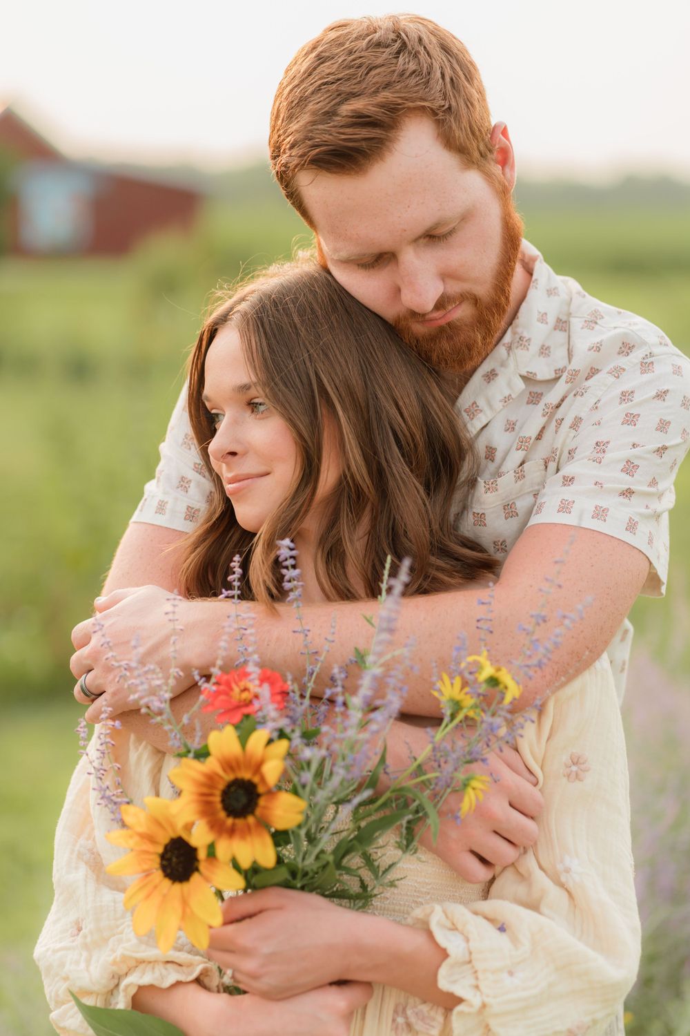 A romantic couple embraces in a field at sunset while holding a bouquet of sunflowers and wildflowers.