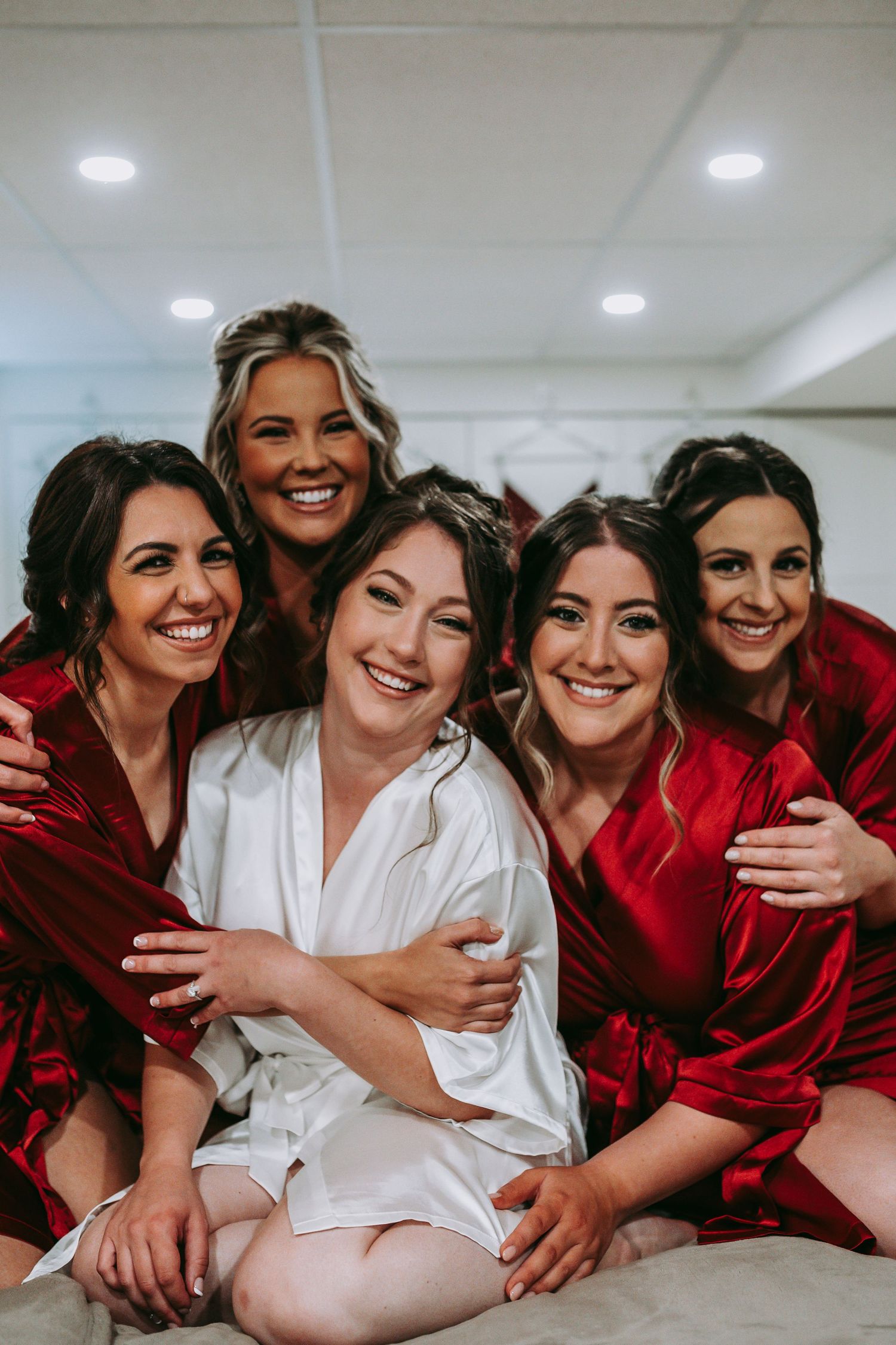 Wedding party in red silk robes and white robe embrace while sharing a candid moment together.