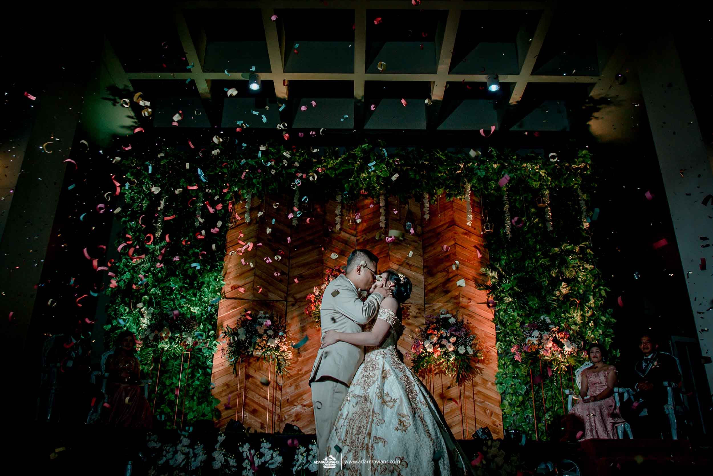 A romantic wedding scene with dramatic lighting, decorative vines, and falling confetti in front of a rustic wall.