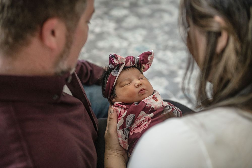 A peaceful newborn wearing a floral headband rests between two caring figures.