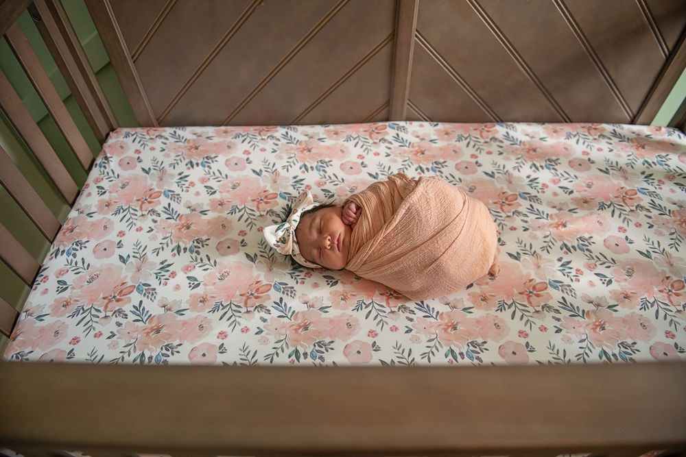A sleeping newborn wrapped in peachy pink swaddle on floral bedding in a mint green crib.