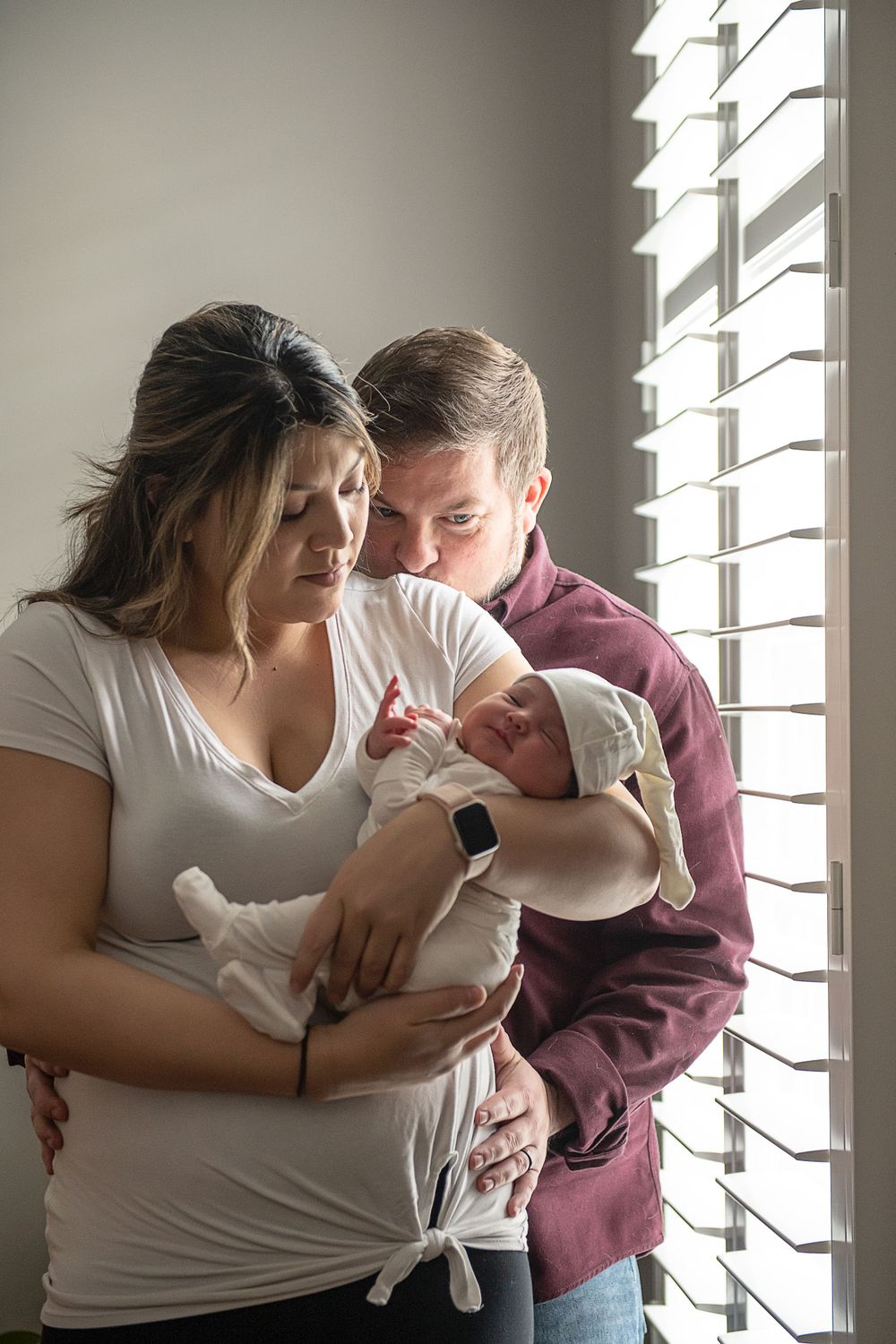 A tender moment by the window showing affection to a swaddled infant in natural light.