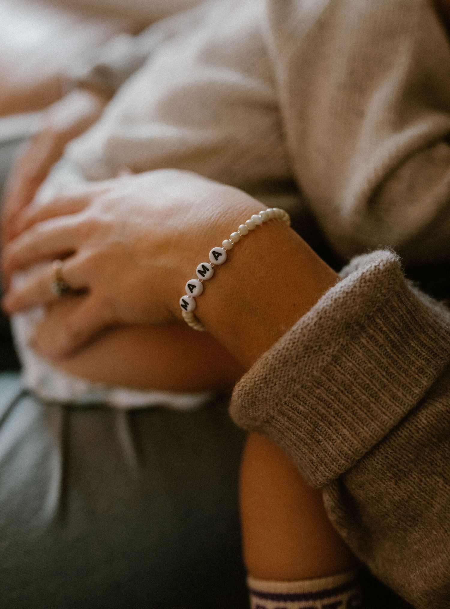 Close-up view of a delicate beaded bracelet saying MAMA on someone's wrist with cozy sweater sleeves.