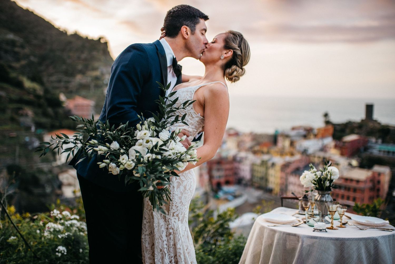 Couple sharing a kiss while bride holds a bouquet of florals with the view from Cinque Terre, Italy in the background as the sunrises.