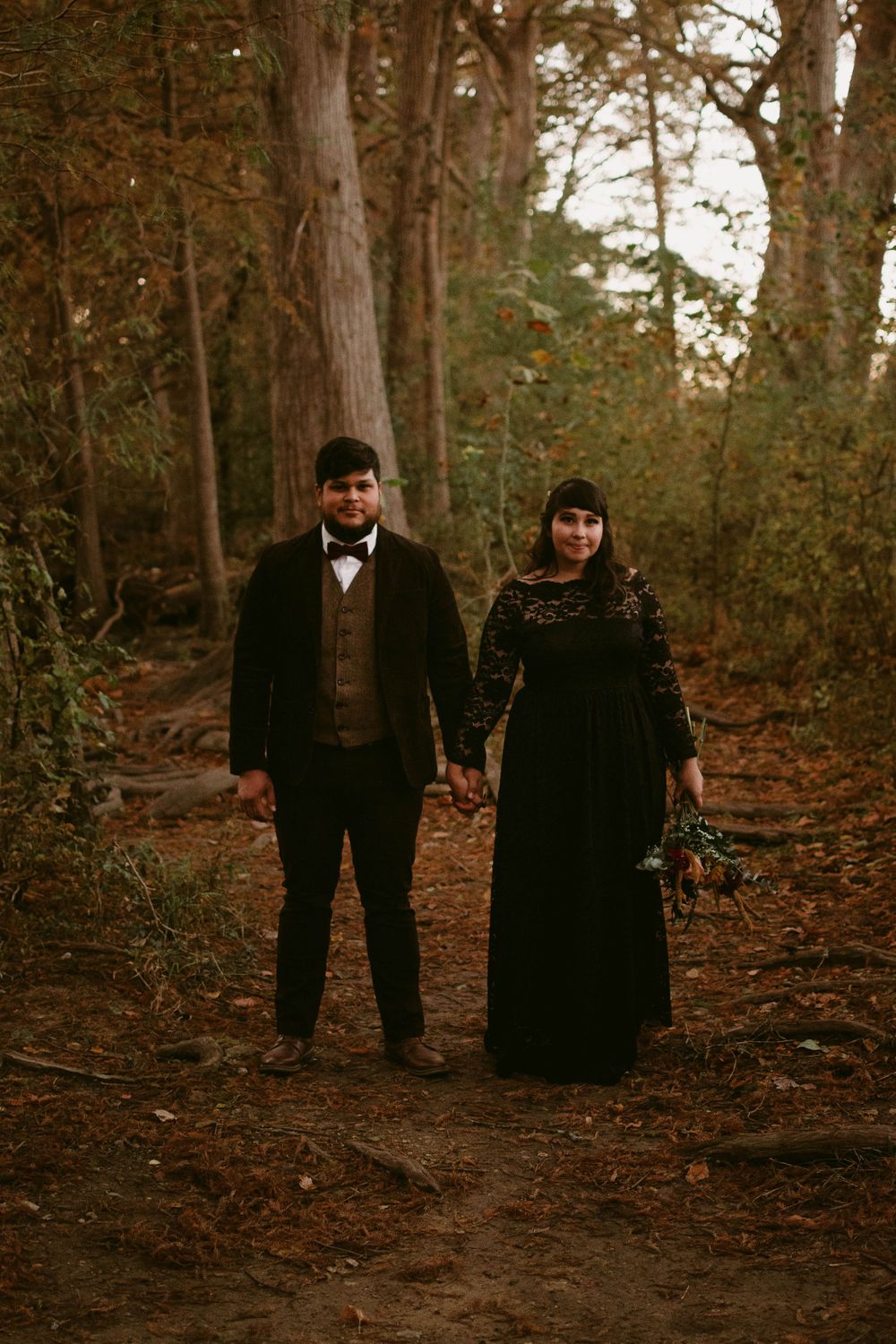 A gothic-styled couple stands together on a forest path wearing formal black attire.