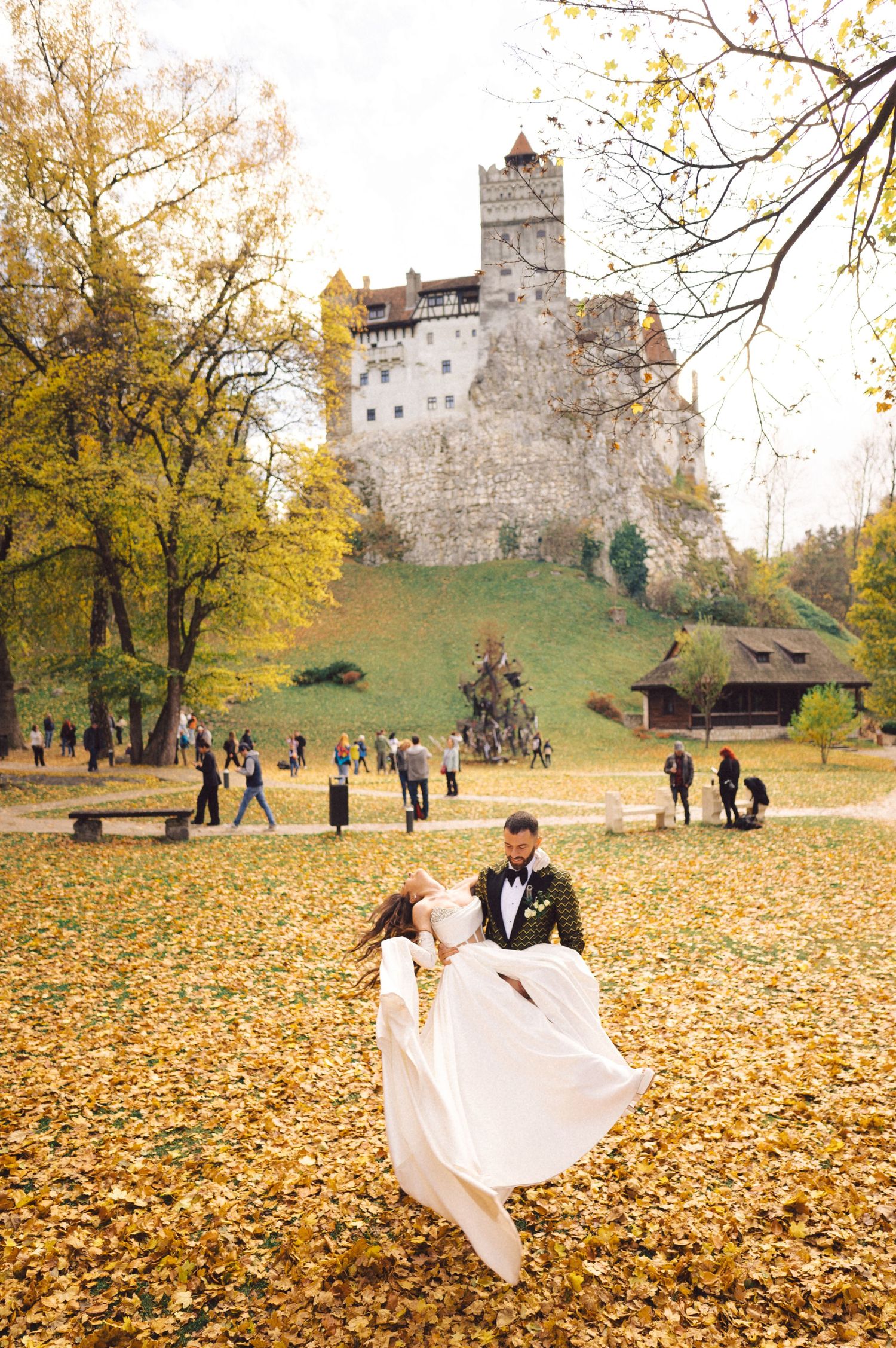 Wedding couple dancing among fallen leaves in front of Bran Castle during autumn.
